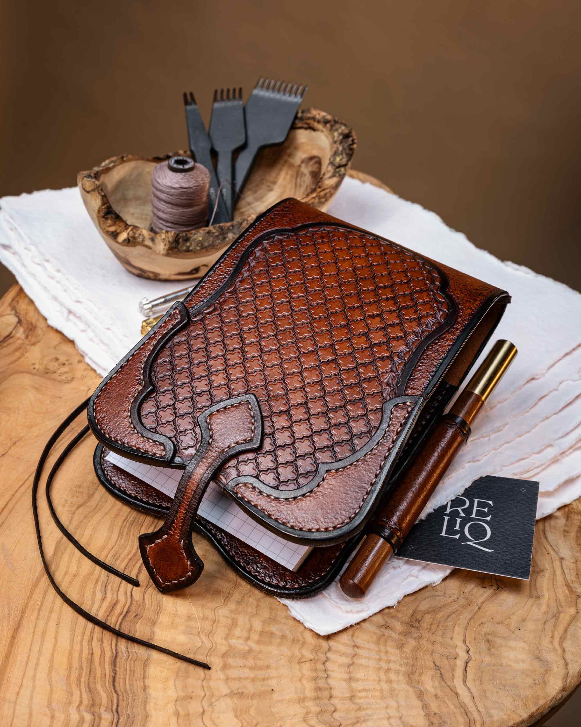 Hand-tooled Italian leather journal cover with the front magnetic closure strap unfastened, resting next to a custom pen and a Reliq brand card.