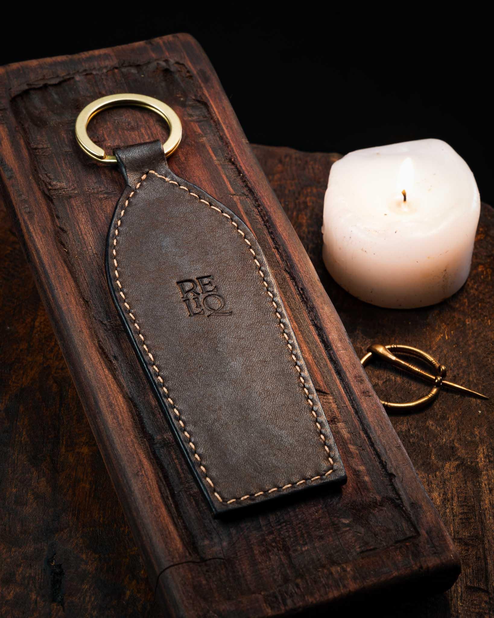 Back view of a handmade medieval leather keyring showcasing smooth tanned leather, detailed edge stitching, and the stamped Reliq workshop logo, resting on a wooden surface next to a lit candle.