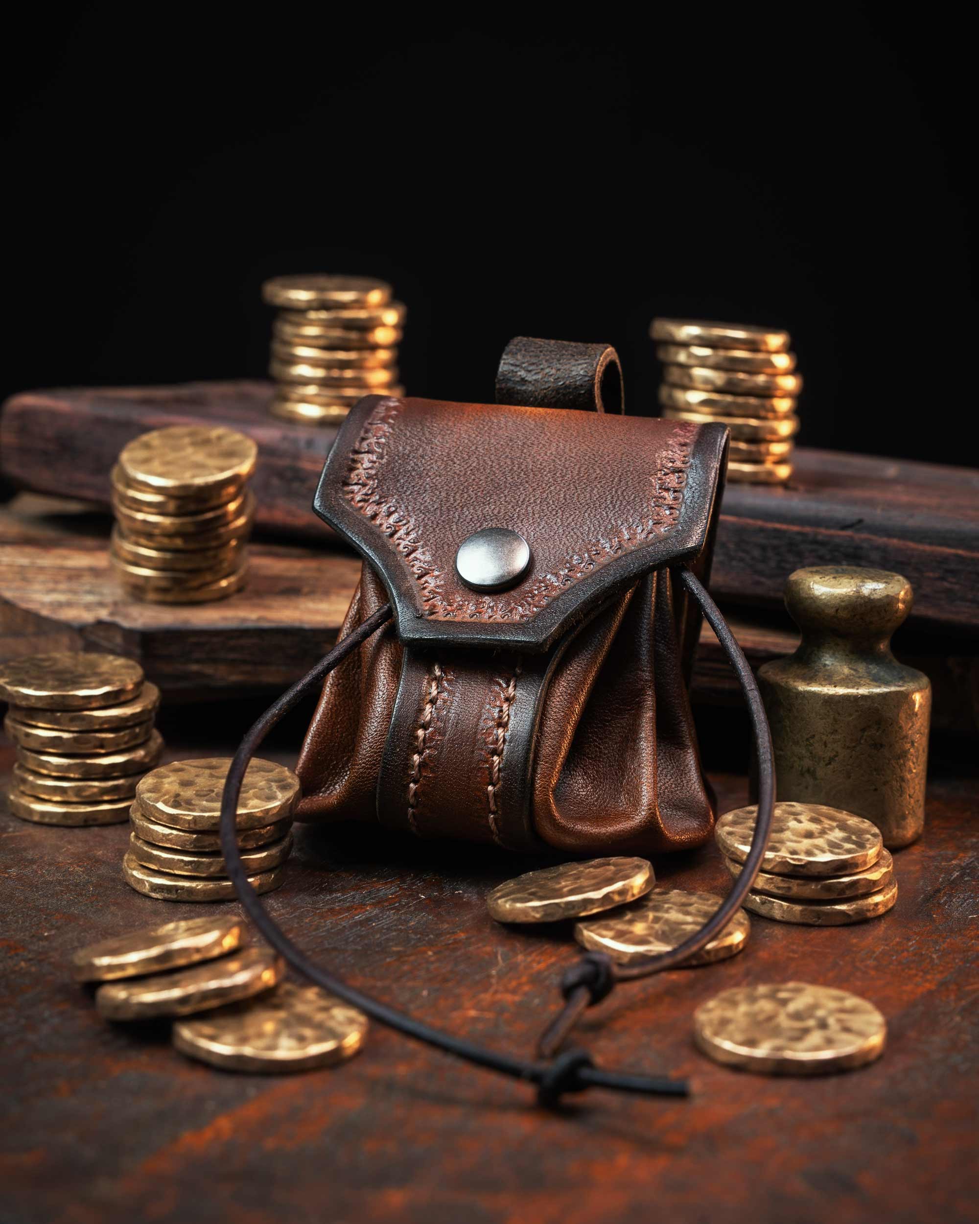 Brown artisan leather belt pouch with a hand-tooled flap, secure snap button, and drawstring bellows design, resting on a wooden surface with metal coins.