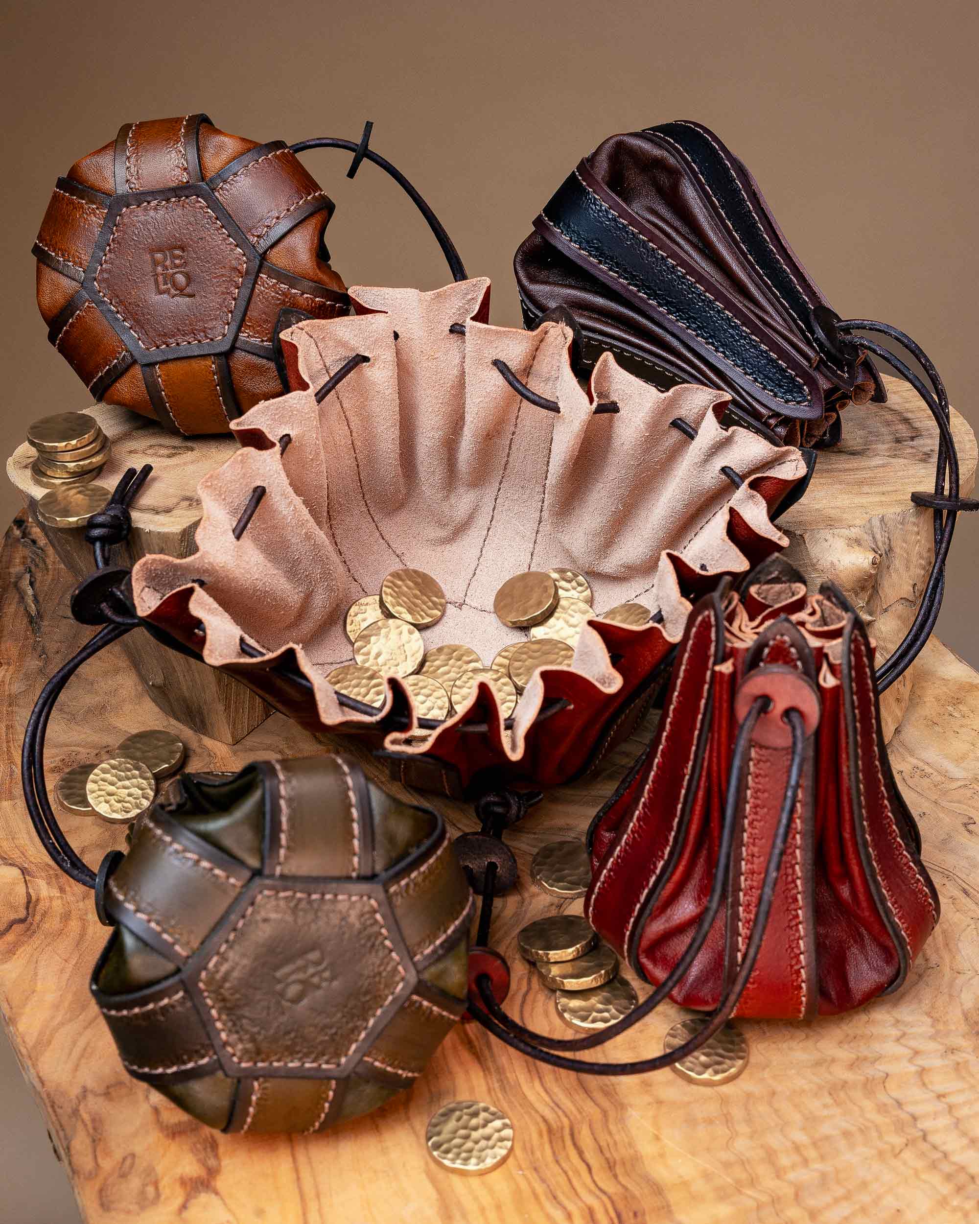 A handcrafted leather drawstring pouch fully opened flat to serve as a catch-all dice tray holding metal coins, surrounded by closed pouches showing the stamped Reliq leather logo.