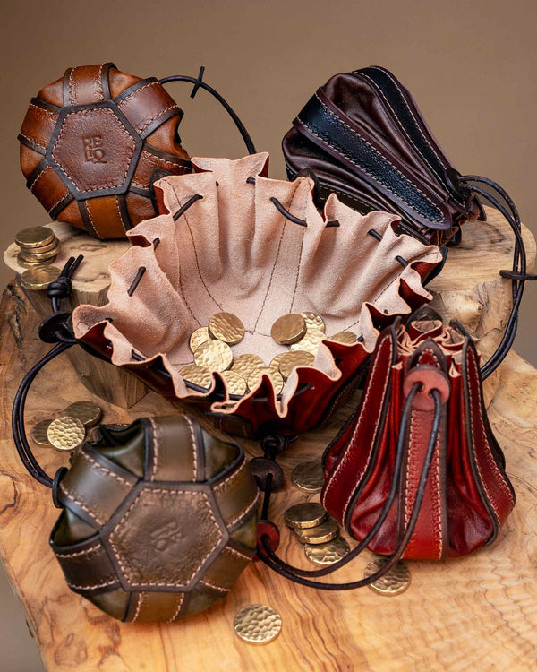 A handcrafted leather drawstring pouch fully opened flat to serve as a catch-all dice tray holding metal coins, surrounded by closed pouches showing the stamped Reliq leather logo.