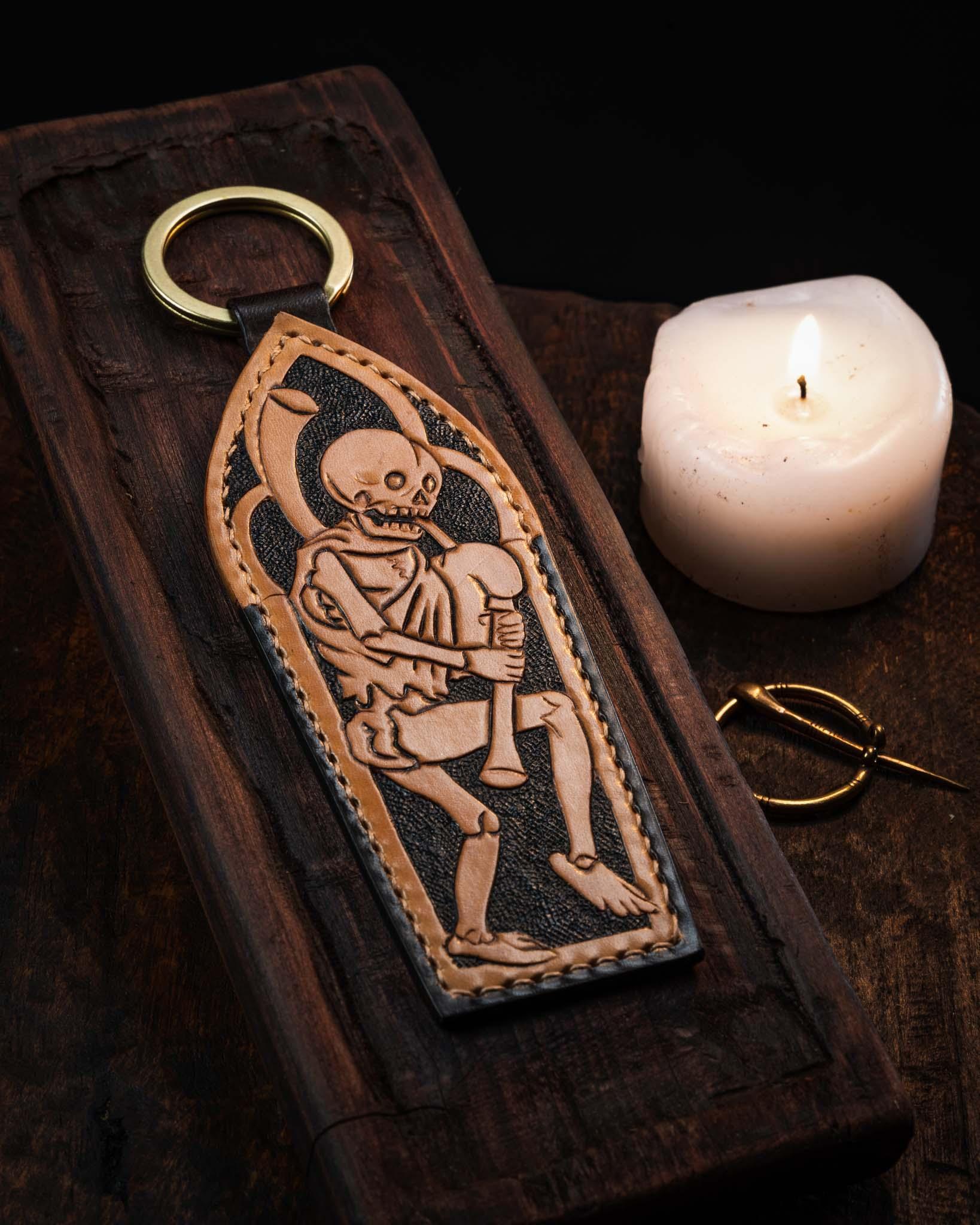 Handcrafted elongated leather keyring featuring a hand-tooled Danse Macabre skeleton playing bagpipes within a gothic arch, set against a textured dark background with a solid brass ring.
