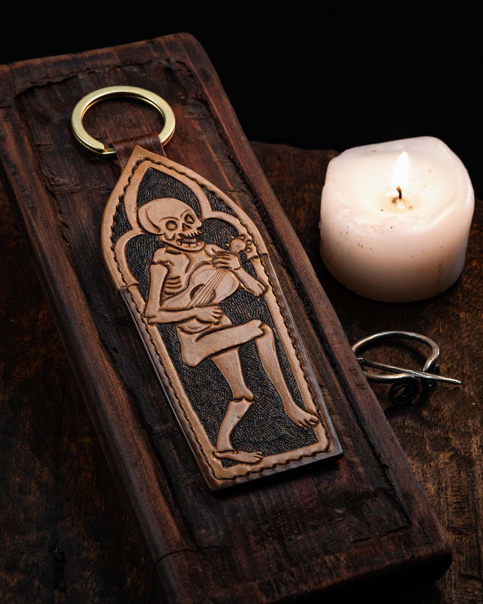 Handcrafted leather keychain featuring a hand-tooled Danse Macabre skeleton playing a lute within a gothic arch, set against a textured dark background with a solid brass ring.