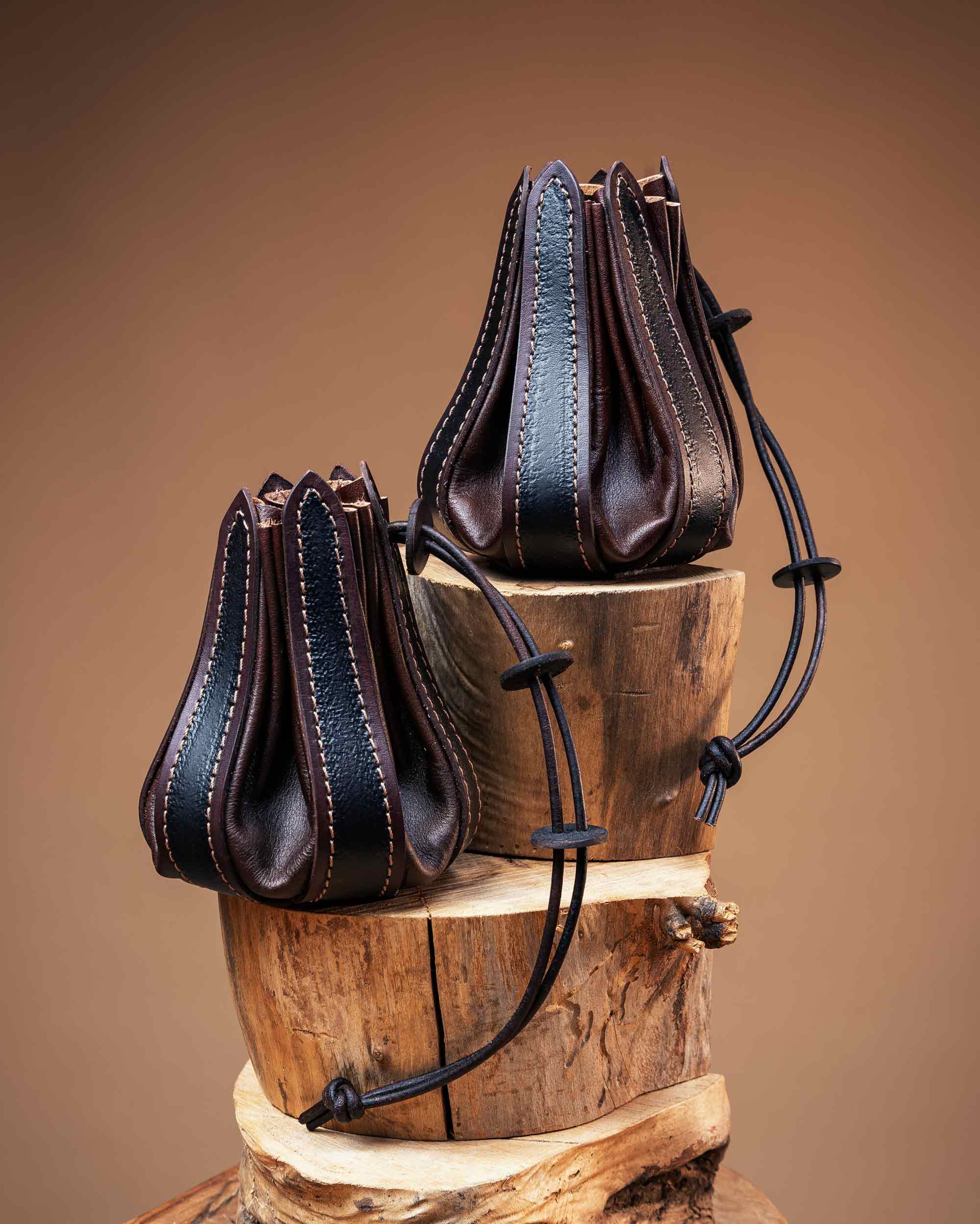 Pair of dark brown leather drawstring pouches with black textured panels and detailed stitching, resting on wooden logs.