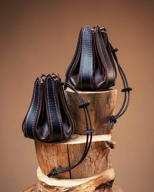 Pair of dark brown leather drawstring pouches with black textured panels and detailed stitching, resting on wooden logs.