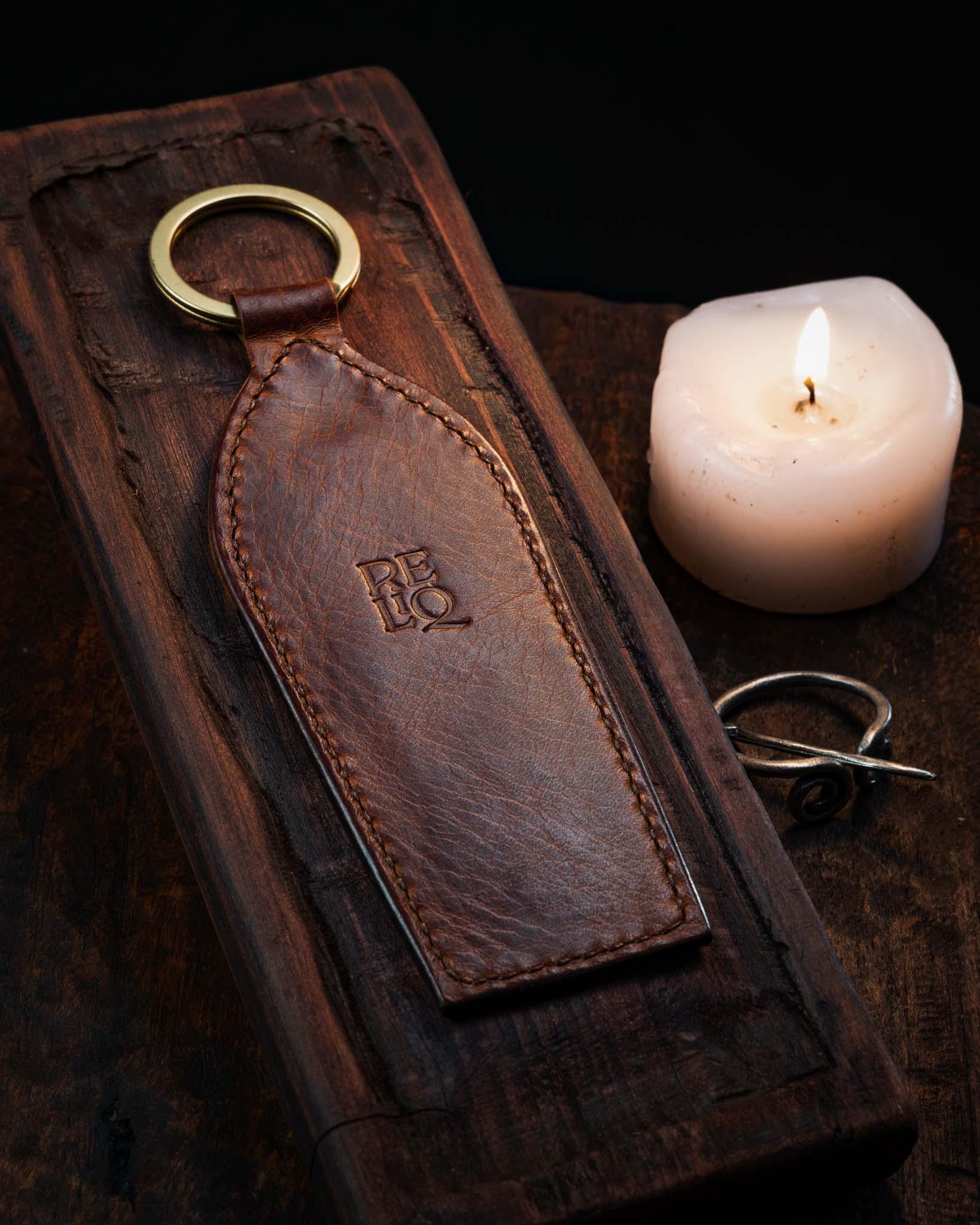 Back view of a handmade elongated leather keychain showcasing smooth tanned leather, detailed edge stitching, and the stamped Reliq workshop logo resting on dark wood next to a lit candle.
