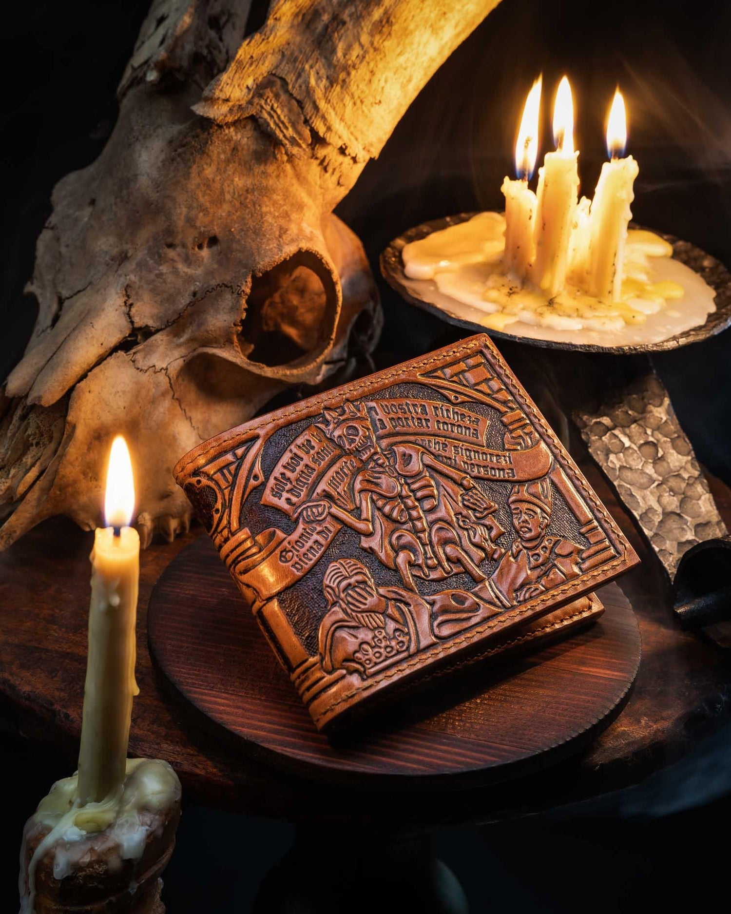 Handcrafted leather bifold wallet featuring an intricate hand-tooled Danse Macabre motif, displayed in a dark setting with lit candles and an animal skull.