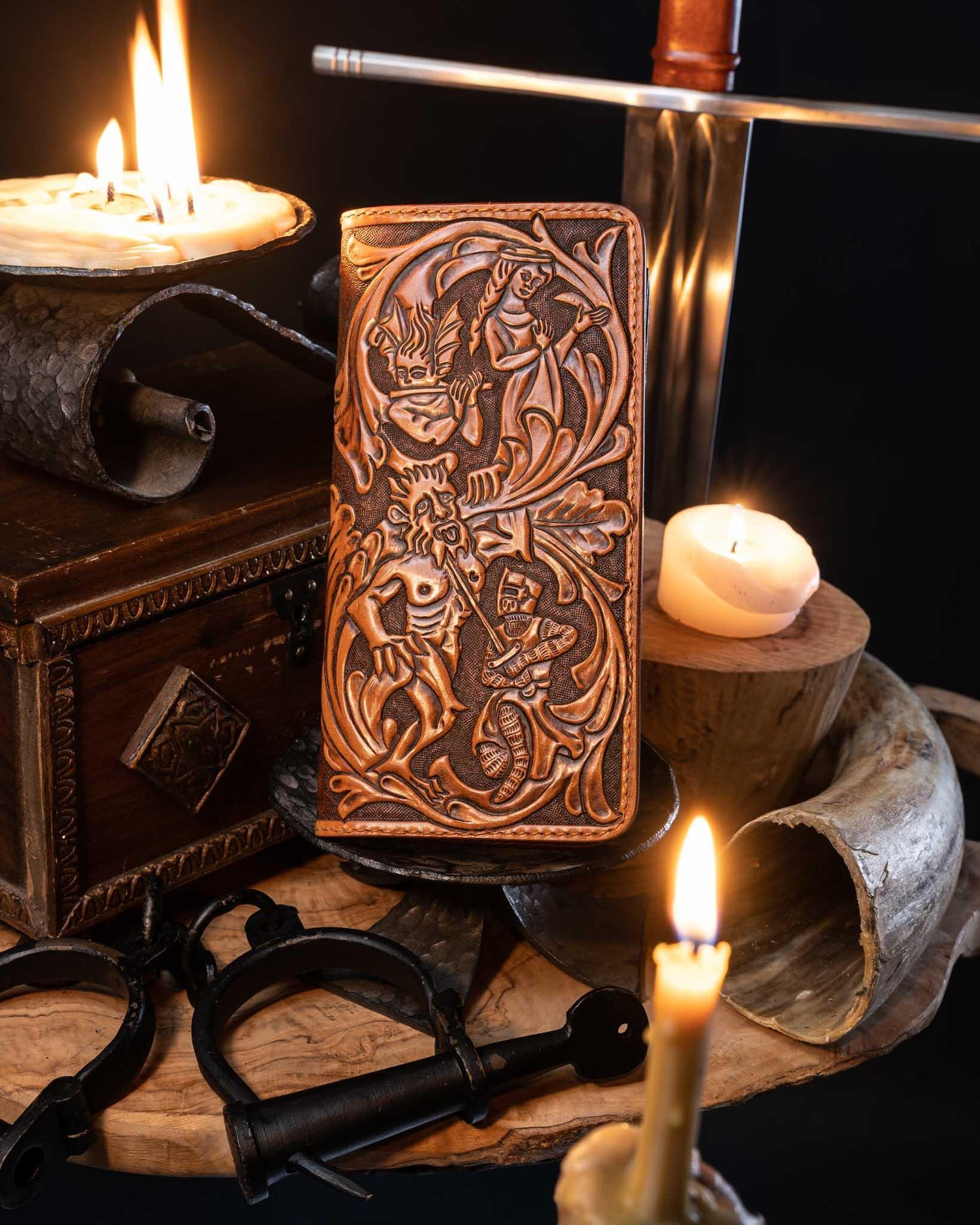 Handcrafted leather long wallet featuring intricate hand-tooled medieval figures inspired by the Huon de Bordeaux epic, displayed alongside a sword and lit candles.