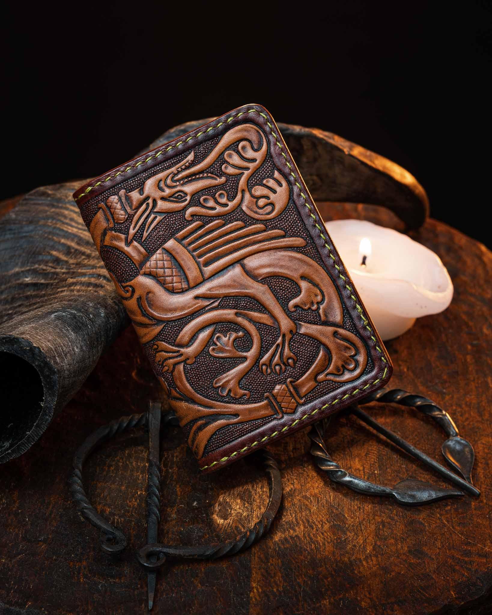 Handcrafted leather card holder featuring an intricate hand-tooled medieval dragon design, resting on a rustic wooden surface next to an iron brooch and a lit candle.
