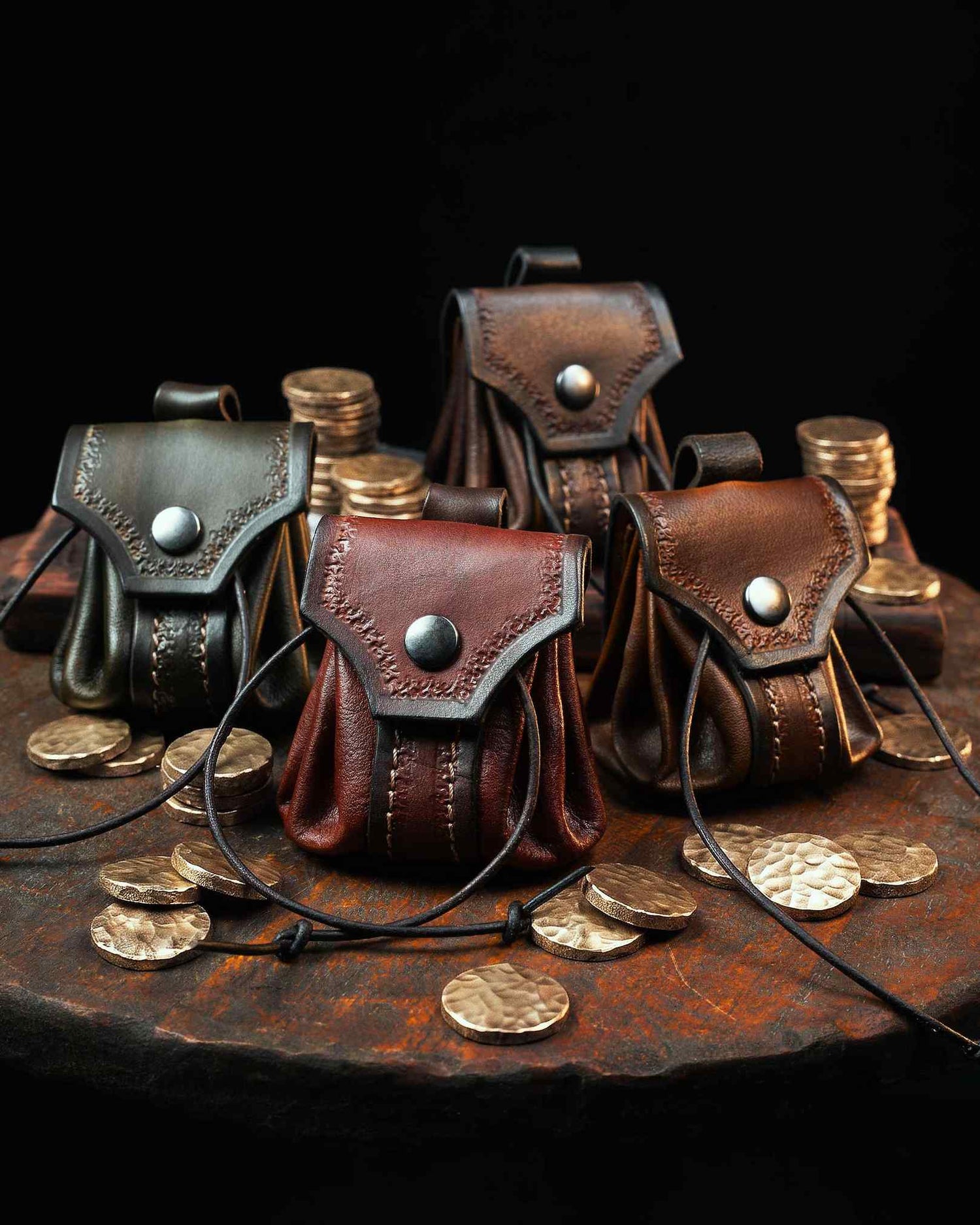 Collection of four handcrafted leather belt pouches in green, red, brown, and dark brown, featuring hand-tooled flaps and snap closures, surrounded by brass coins.