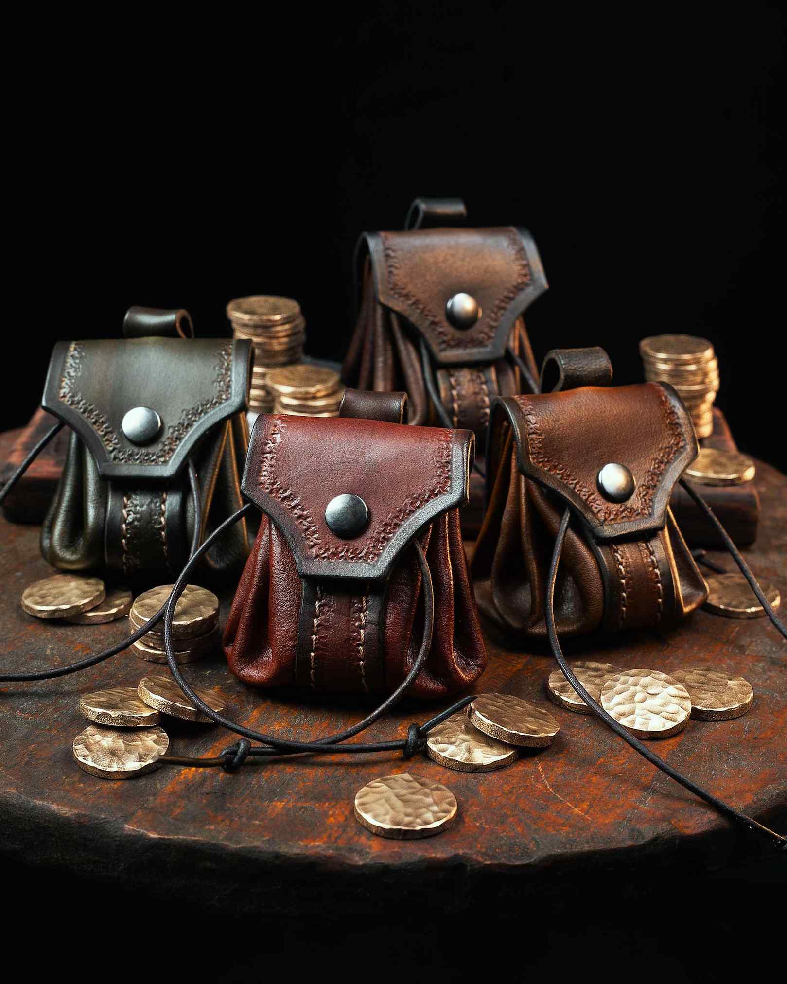 Collection of four handcrafted leather belt pouches in green, red, brown, and dark brown, featuring hand-tooled flaps and snap closures, surrounded by brass coins.