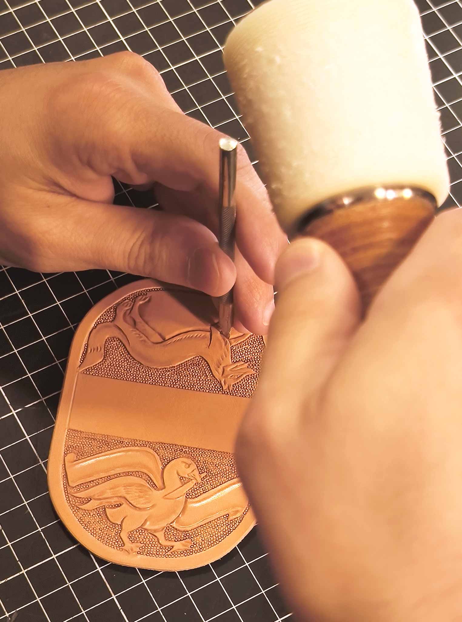 Close-up of Michele Belvedere hand-tooling a heraldic design on vegetable-tanned leather for a custom sword rainguard. Background being worked with precision tools.
