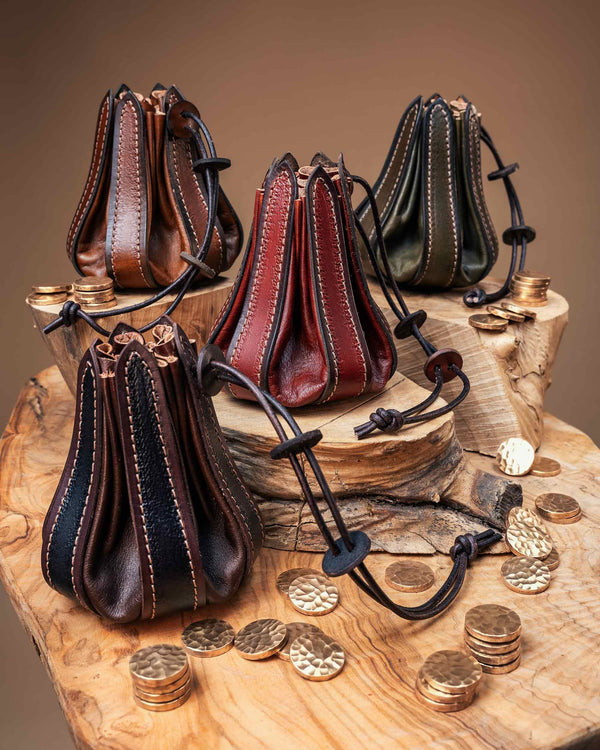 Four handcrafted leather bellows pouches in brown, red, green, and black, arranged on a rustic wooden slab next to brass metal coins.