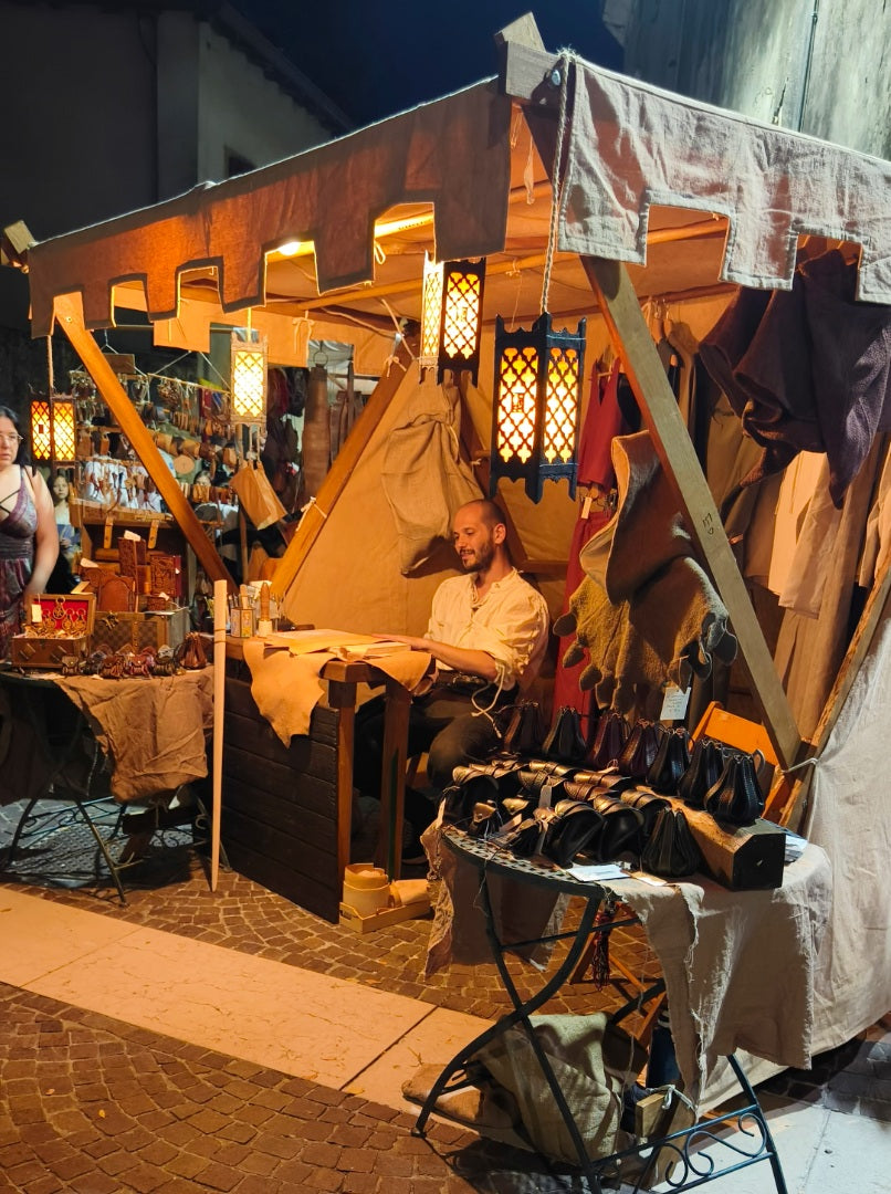 Night view of a historical reenactment tent where a craftsman is showing the steps of leather working live to the public, surrounded by an exhibition of handmade leather accessories