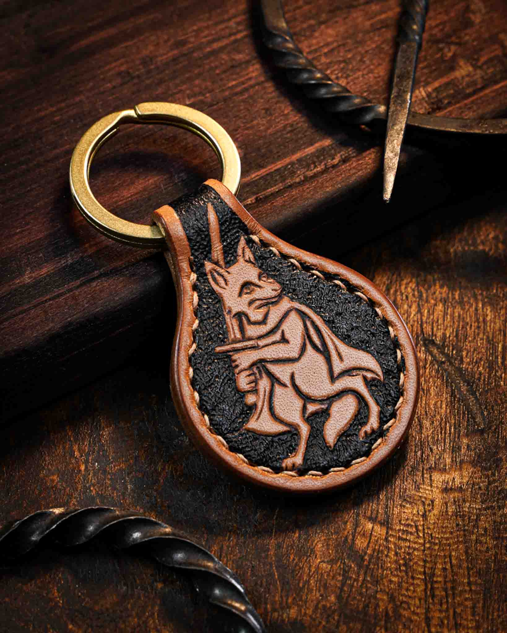 Handcrafted leather keyring featuring a hand-tooled medieval marginalia fox warrior design with a textured dark background, finished with stitched edges and a solid brass ring.