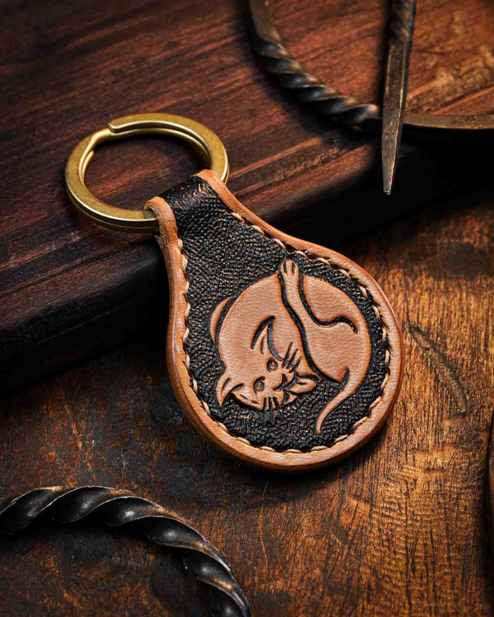 Handcrafted leather keyring featuring a hand-tooled medieval marginalia cat design with a textured dark background, finished with stitched edges and a solid brass ring.