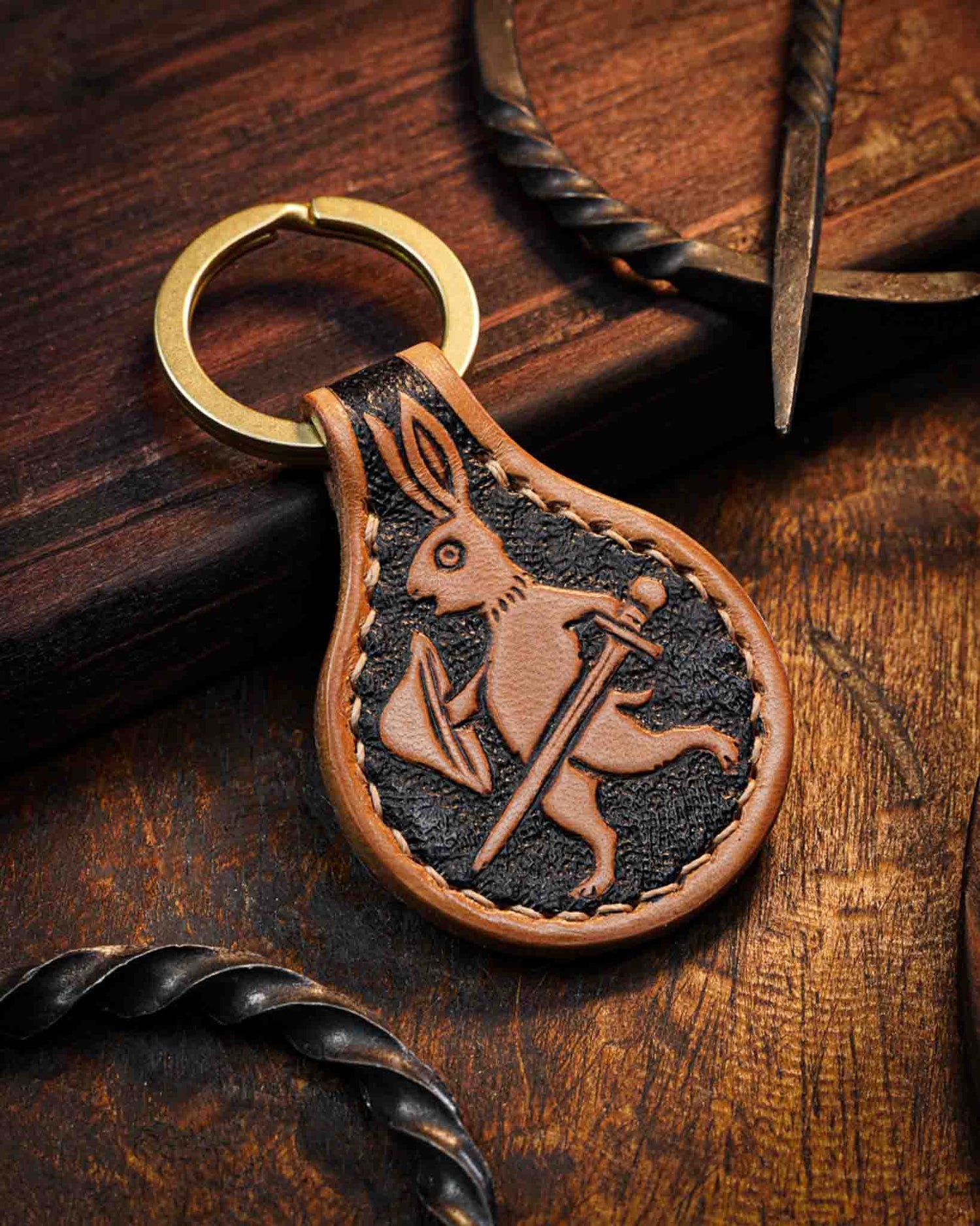 Handcrafted leather keyring featuring a hand-tooled medieval marginalia rabbit armed with a sword, set against a textured dark background, finished with stitched edges and a solid brass ring.