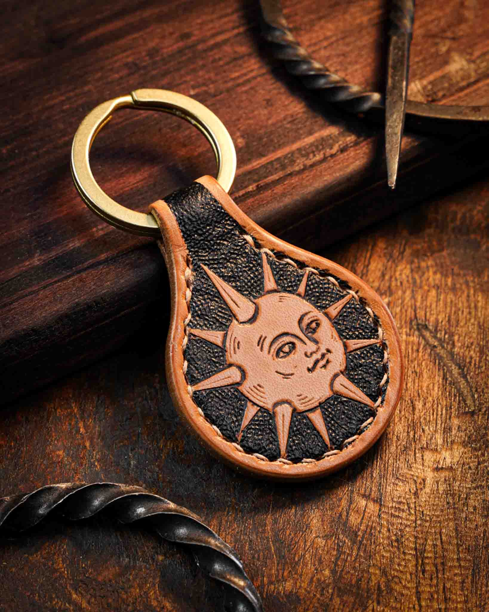 Handcrafted leather keyring featuring a hand-tooled medieval sun talisman design with a face, set against a textured dark background, finished with stitched edges and a solid brass ring.
