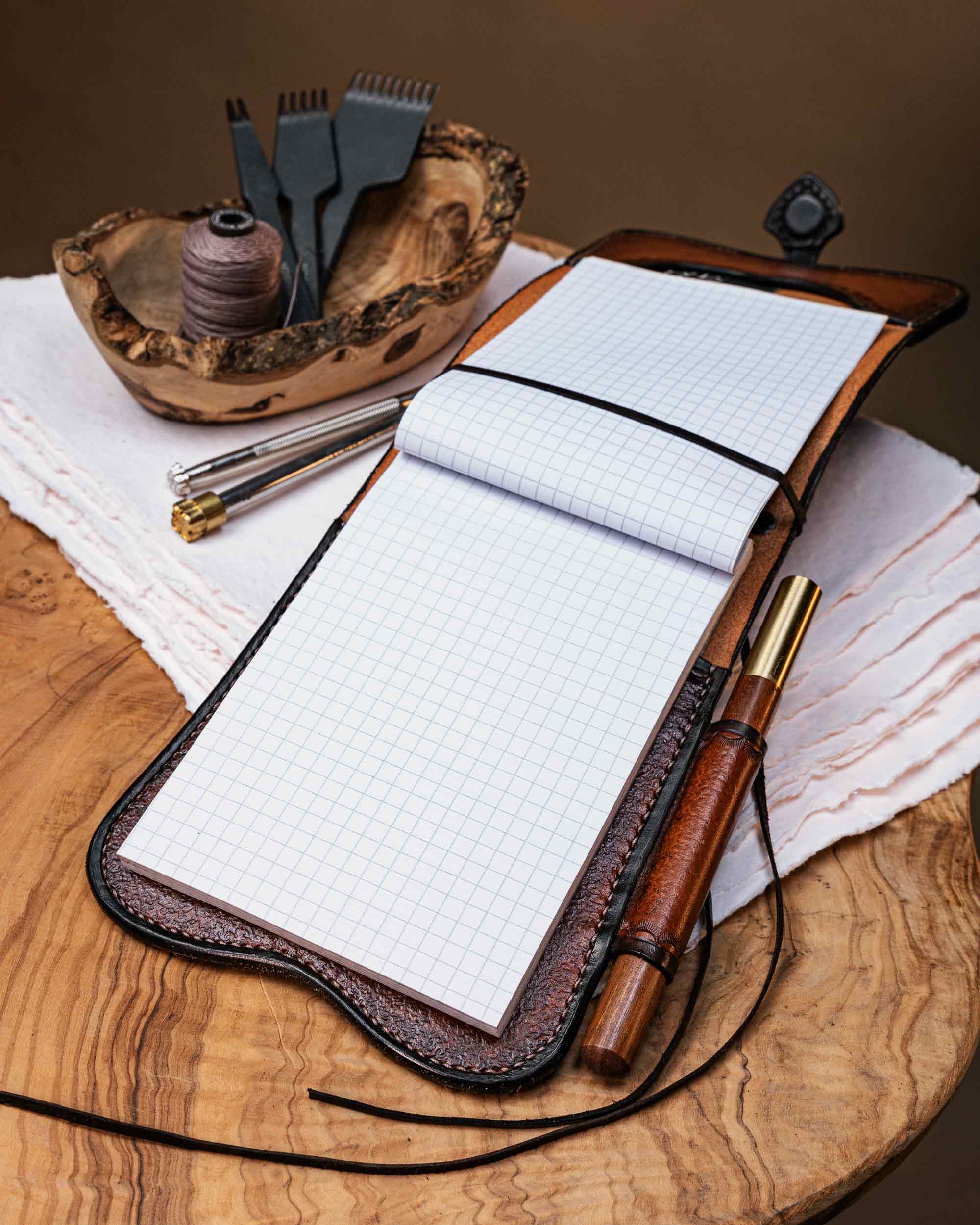 Open handcrafted A6 leather journal cover displaying a refillable graph paper notebook secured with an kangaroo leather band, styled with a matching pen and leathercraft tools.