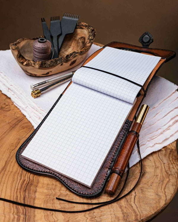 Open handcrafted A6 leather journal cover displaying a refillable graph paper notebook secured with an kangaroo leather band, styled with a matching pen and leathercraft tools.