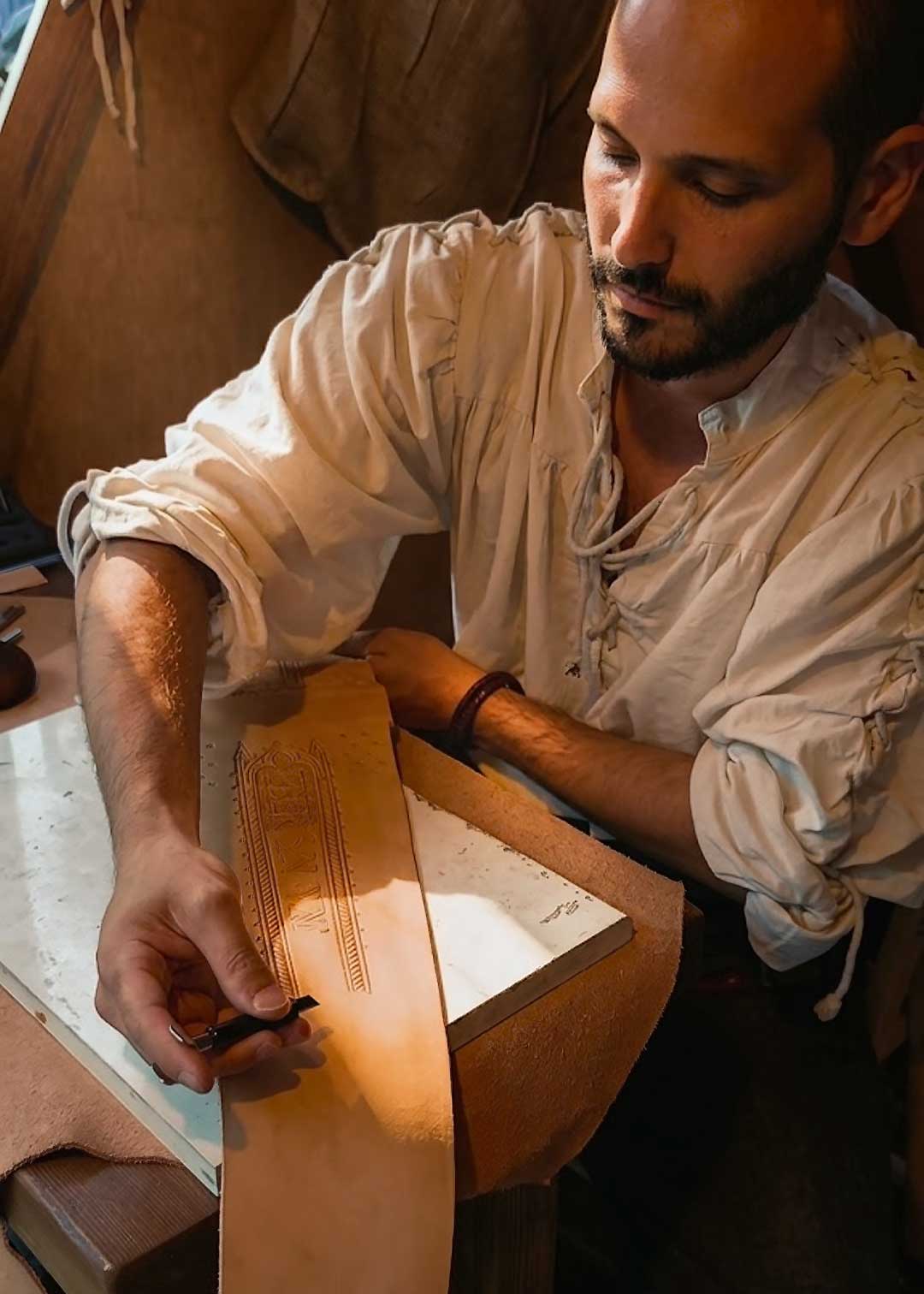 Michele Belvedere, artisan of Reliq Leather, dressed in medieval clothing, focusing on cut a complex hand-tooling pattern onto a new leather sword scabbard. A dramatic shaft of sunlight from an upper opening illuminates his hands and the precision swivel knife, while the rest of the dim, canvas-sack workshop is in warm, soft shadow. Marble block and leather scraps are on the wooden workbench. This live creation took place at a historical re-enactment event.