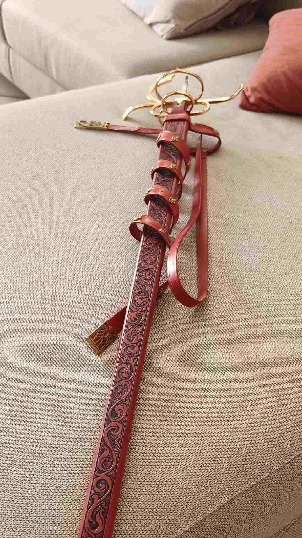 Full-length view of a custom red leather scabbard featuring intricate hand-tooled floral patterns, wrapped with a matching leather sword belt and brass hardware, sheathing a complex-hilted historical sword
