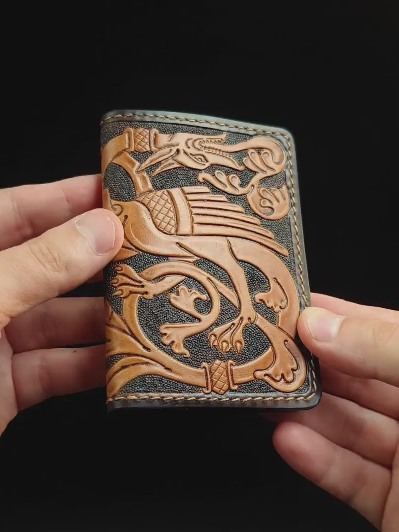Video demonstration of a hand-tooled medieval leather card holder. Hands display the carved marginalia dragon on the front, turn it to reveal foliate scrollwork on the back, and open the artisan wallet to show the two-tone interior, sliding a card into the Reliq Leather branded pocket.