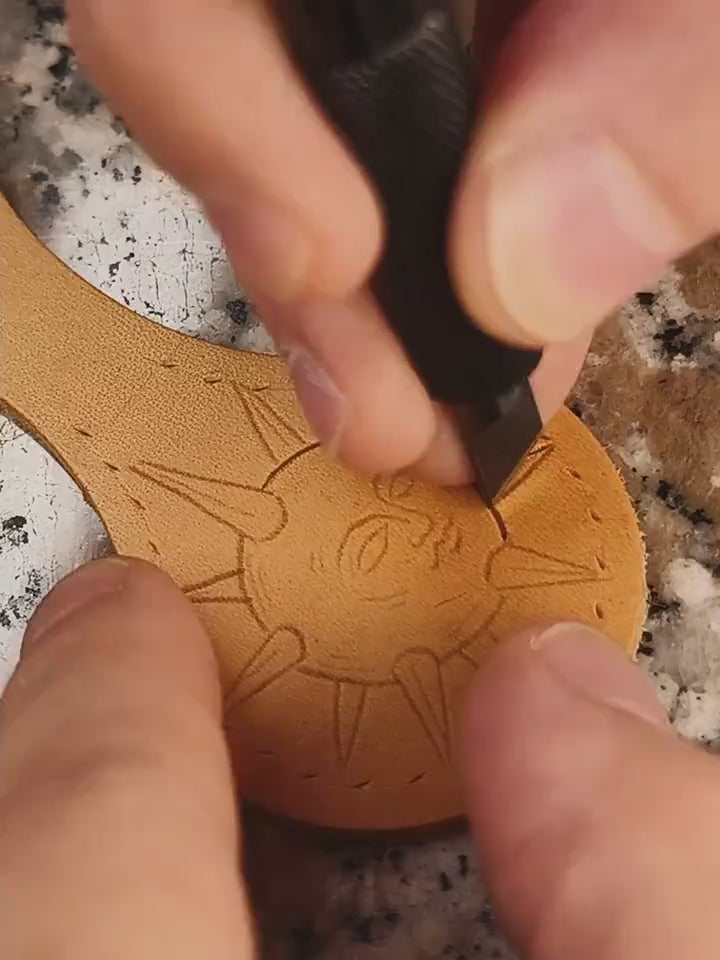 Making of the medieval sun talisman leather keyring: close-up of the hand-tooling process using a swivel knife to cut the design, followed by a beveler and backgrounding tools to add depth and texture.