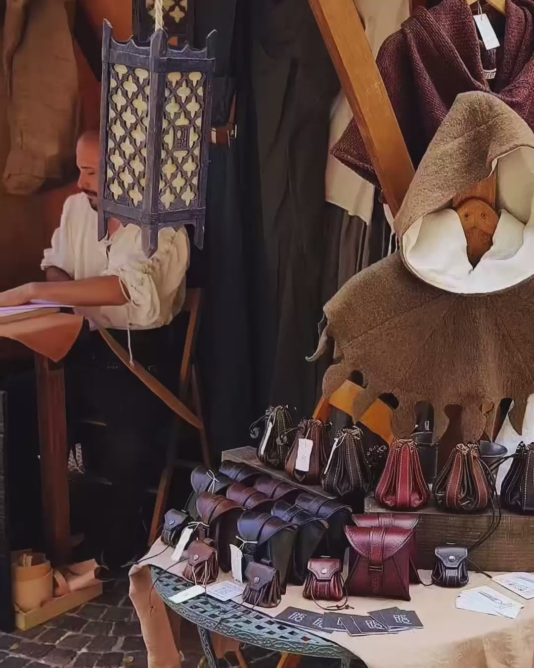 Video showcasing the leather artisan working at a medieval-themed display filled with handcrafted bellows pouches, historical garments, and custom leather accessories.