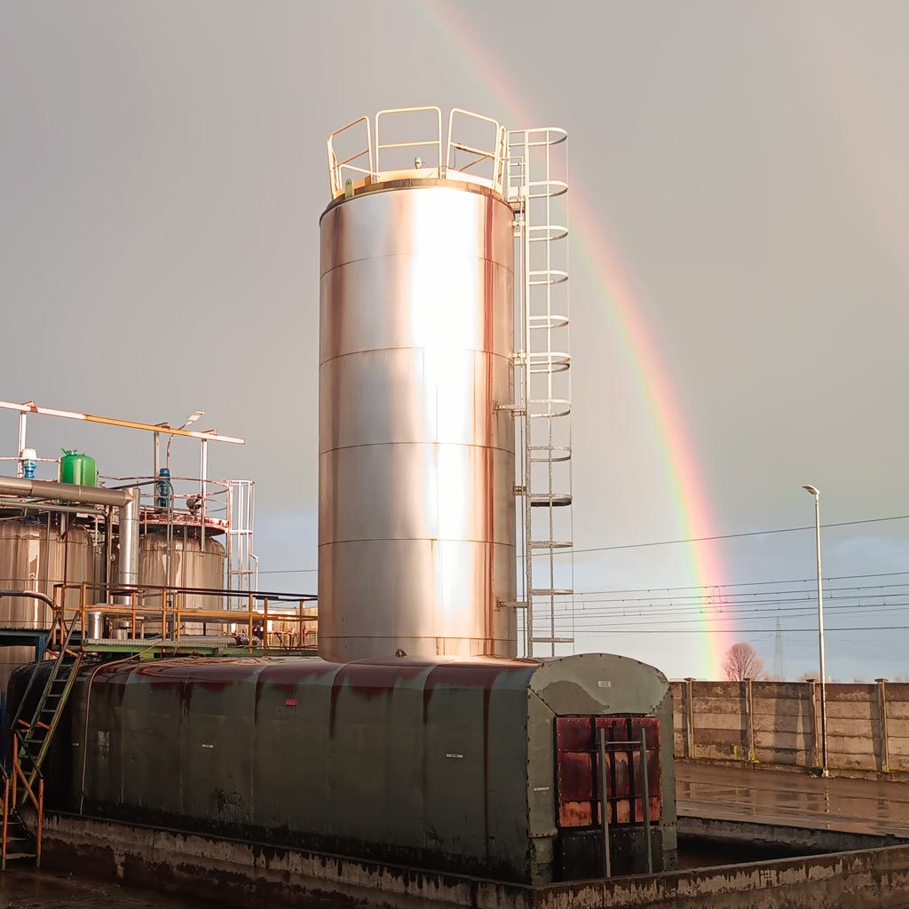Industrial steel silos under a grey sky with a rainbow, serving as the signal for the final leap into the Reliq quest in 2025.