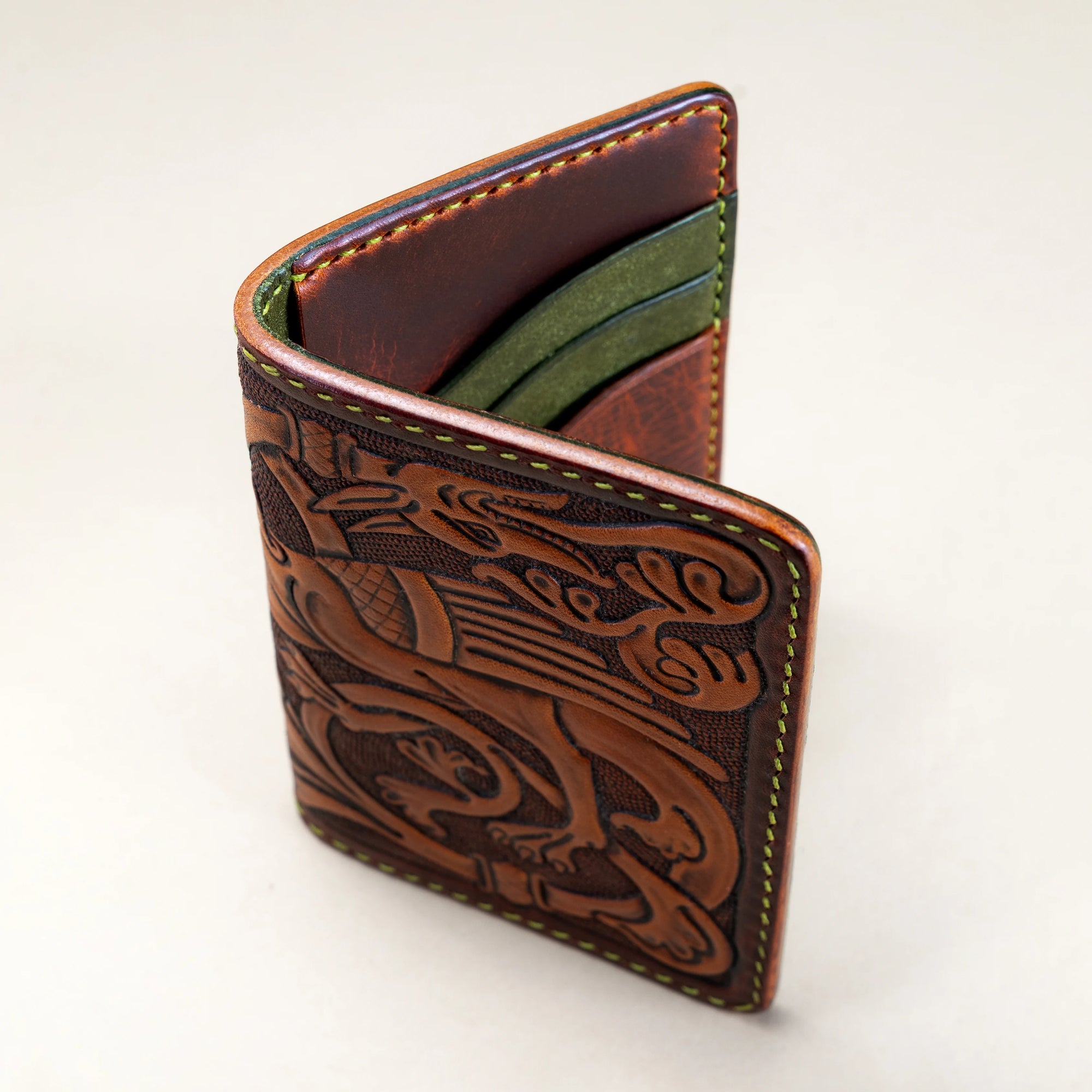 Angled view of the Western Dragon leather card holder wallet by Reliq, showing hand-tooled dragon design and contrasting mahogany and green tones with olive linen stitching.
